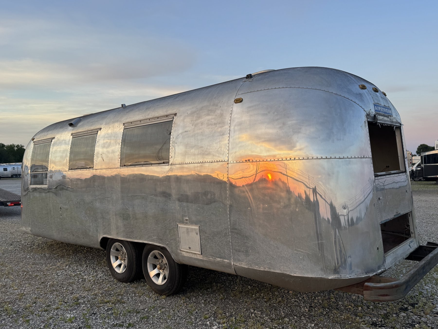 The polished Silver Bullet Airstream parked on gravel at the Airstream factory in Jackson Center, Ohio, at sunset, with the sky reflected in its aluminum side and the service window opening visible.