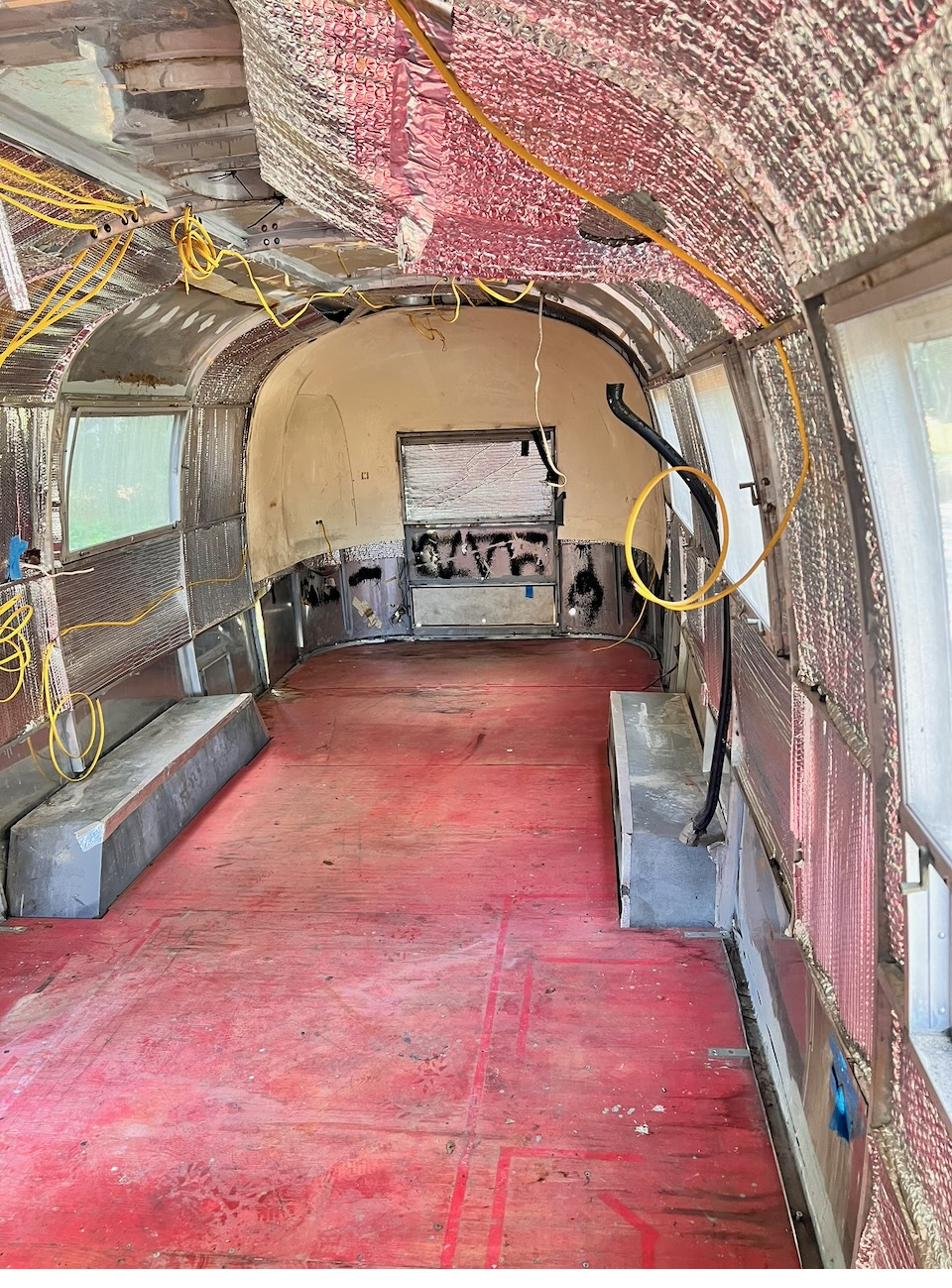Interior of the Silver Bullet Airstream stripped to the bare aluminum shell, looking down the length toward the front, with a red painted plywood floor and exposed wiring.